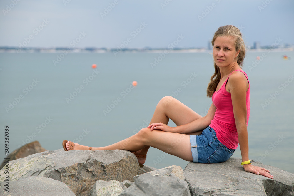 Young woman in summer clothes sitting on big boulders on sea shore.