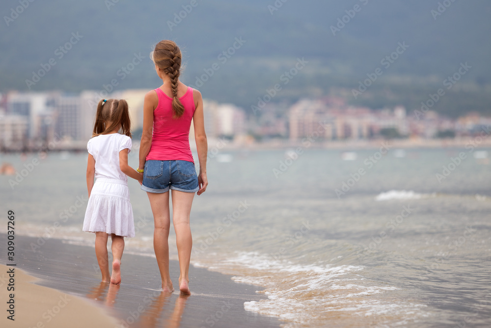Mother and daughter girl walking together on sand beach in sea water in summer with bare feet in warm ocean waves. Mother and daughter girl walking together on sand beach in sea water in summer with bare feet in warm ocean waves.