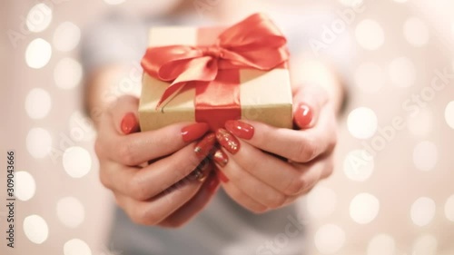 Unrecognizable young woman gives a gift box with red ribbon to camera on blurred lights.