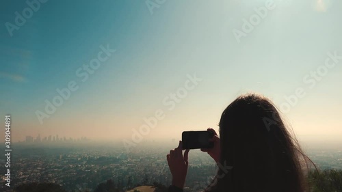 4K. Young woman photographs downtown Los Angeles in the distance at the Griffith Park Observatory. The fog makes the buildings look more interesting. Woman makes photos of the Los Angeles skyline. 