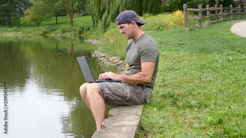 Man sitting by a lake, river, pond, working on a laptop computer, typing.