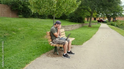 Middle aged young man sitting on a bench in the park recieves bad news and is stressed while working on his laptop computer.