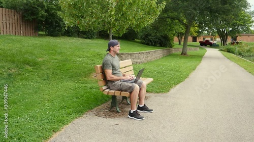 Man sitting on a bench in a park, thinking and working and typing on a laptop computer.