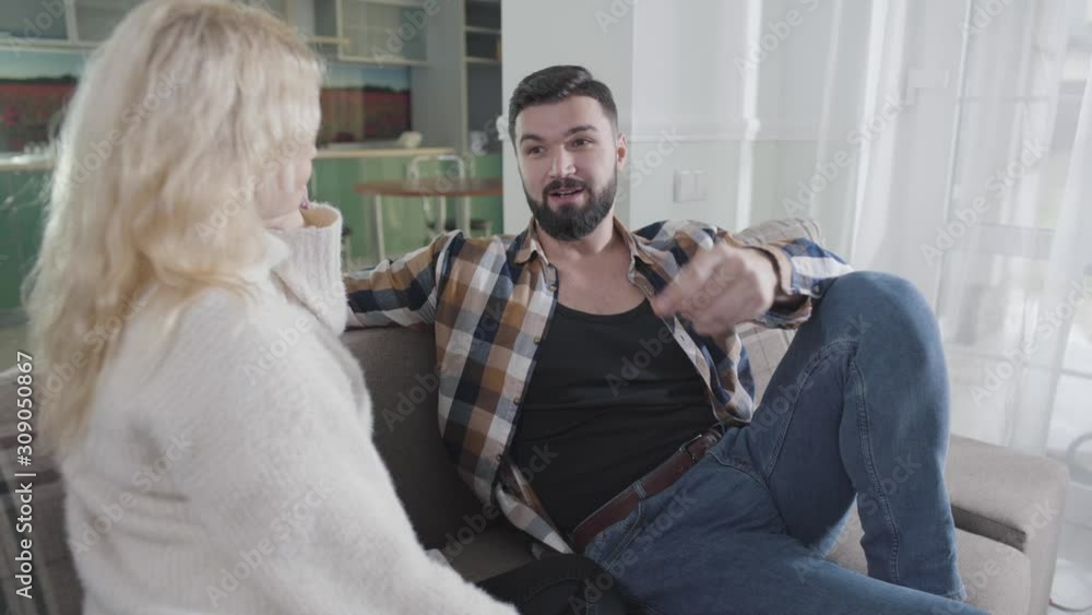 Close-up of handsome Caucasian man with black hair and beard sitting on couch and talking to blond woman. Guy in casual clothes spending free time with wife or girlfriend at home. Leisure, weekends. Close-up of handsome Caucasian man with black hair and beard sitting on couch and talking to blond woman. Guy in casual clothes spending free time with wife or girlfriend at home. Leisure, weekends.