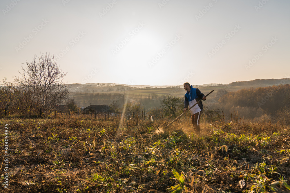 Young handsome man mows dry grass, amazing autumn lanscape with sunset on the background
