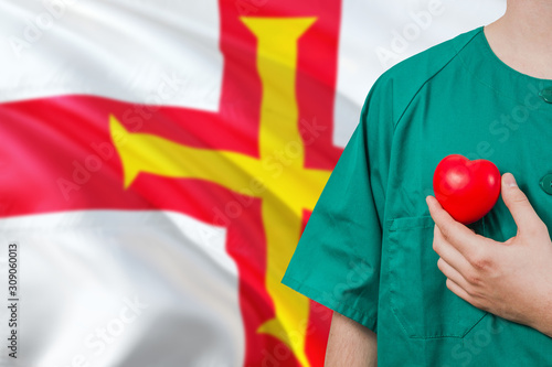 Guernsey veterinary clinic concept. Veterinarian is holding plastic heart in green uniform on national flag background. Animial love theme. photo