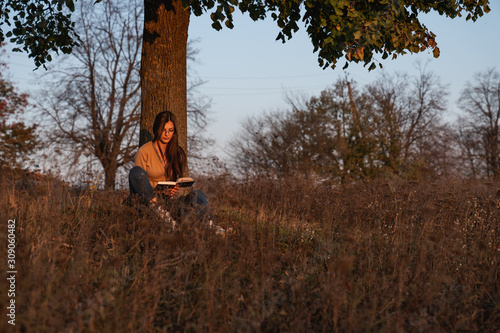 Student female in casual clothes sitting with book under the tree among the meadow