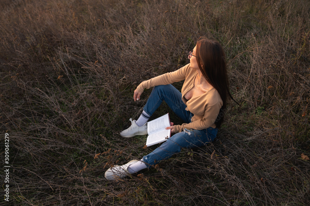 Attractive brunette girl with loose hair dressed in casual clotehs sitting on the groung among the meadow with book in hands and looking away