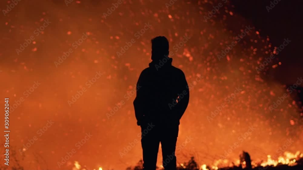Slow motion, pan shot, around a silhouette man standing in front of fire, smoke, ash and sparks, at the Californian wildfires, at night time, in Los Angeles, California, USA