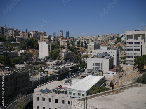 Cityscape of Amman, capitol of Jordan, grey panorama of a modern Arabic city with improvised houses on a hill between few green trees and a tall flag of the country under the blue sky