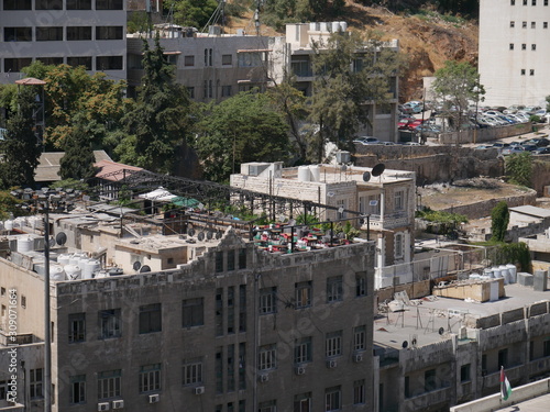Cityscape of Amman  capitol of Jordan  grey panorama of a modern Arabic city with improvised houses on a hill between few green trees and a tall flag of the country under the blue sky