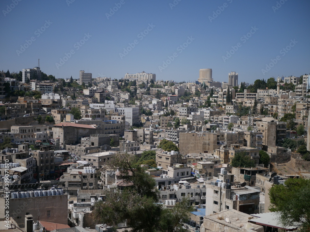 Cityscape of Amman, capitol of Jordan, grey panorama of a modern arabic city with improvised houses on a hill between few green trees under the blue sky