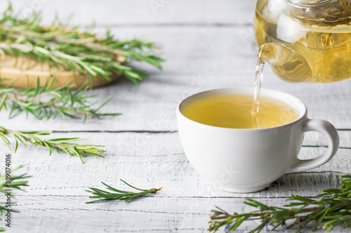 White cup of healthy rosemary tea pouring from teapot with fresh rosemary bunch on white wooden rustic background, winter herbal hot drink concept, salvia rosmarinus photo