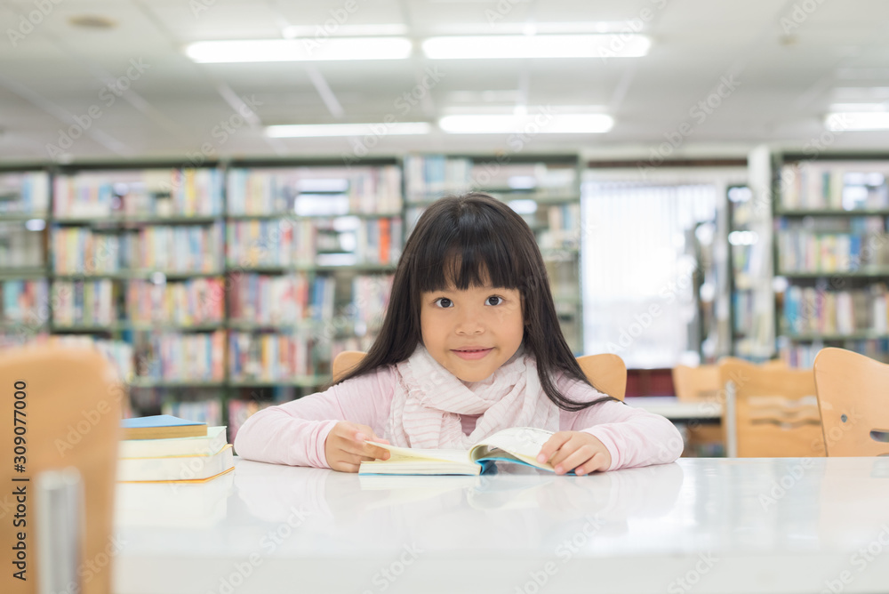 portrait of cute schoolgirl smiling while sitting with stack of books at table in library