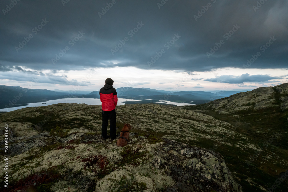 A hiker with red jacket and his dog are standing on a mountain top looking towards lakes and dramatic stormy clouds during blue hour. Hiking and active lifestyle concept.