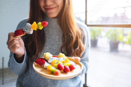 Closeup image of an asian woman holding and eating a fresh mixed fruits on skewers