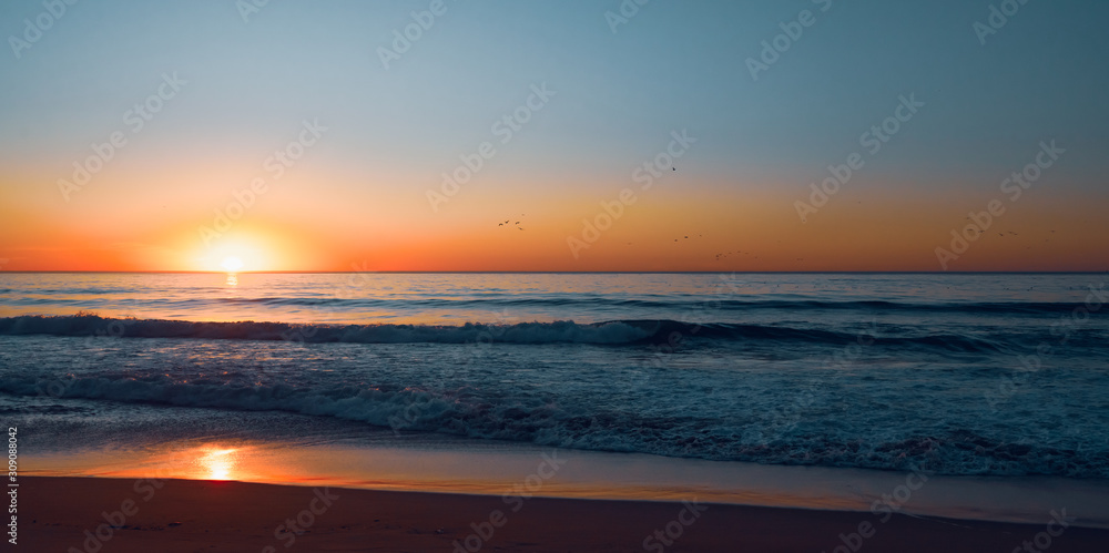 Abstract seascape. Sunset over the ocean. Blue ocean waves, sand beach, colorful sky, and sun setting down the horizon