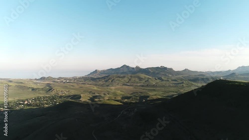 Aerial view of the small settlement in green hills with high mountain chain and blue sea on the background against blue sky. Shot. Beautiful colorful landscape.