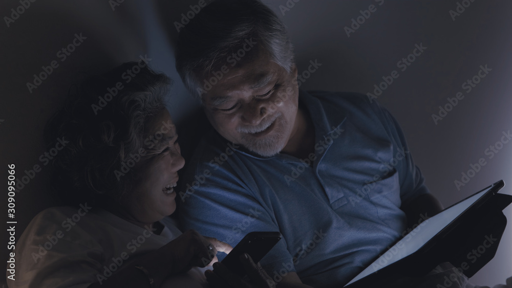 couple lovely senior elderly man and woman, sitting and watching internet TV and happy smile together in the bedroom at night, technology and healthcare, retirement lifestyle concept