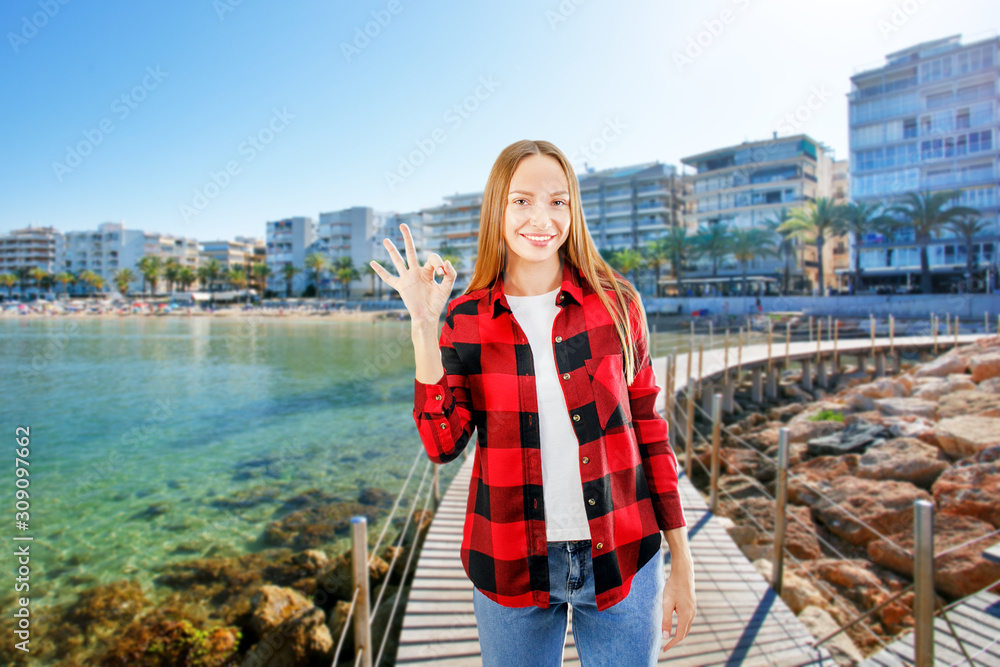 Happy smiling young woman showing ok hand sign against beach background. Travel, tourism and summer holidays concept