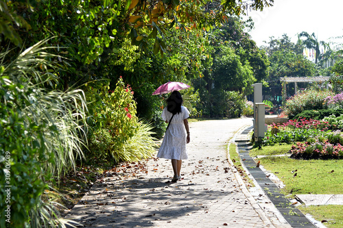 A woman wear white dress hold a pink umbrella walking on street under sunlight and tree leaves fall on the ground, alone on the way.