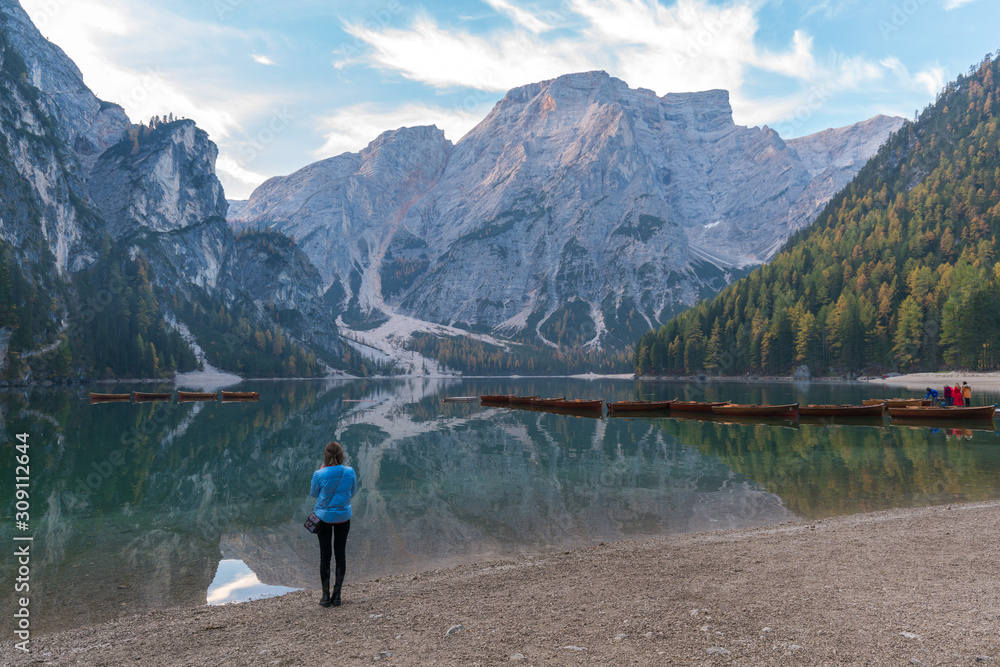 A woman standing beside the lake Braies (Lago di Braies) in a morning with reflection of the mountain peak and boats on the calm lake in Dolomites, Italy