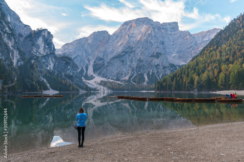 A woman standing beside the lake Braies (Lago di Braies) in a morning with reflection of the mountain peak and boats on the calm lake in Dolomites, Italy