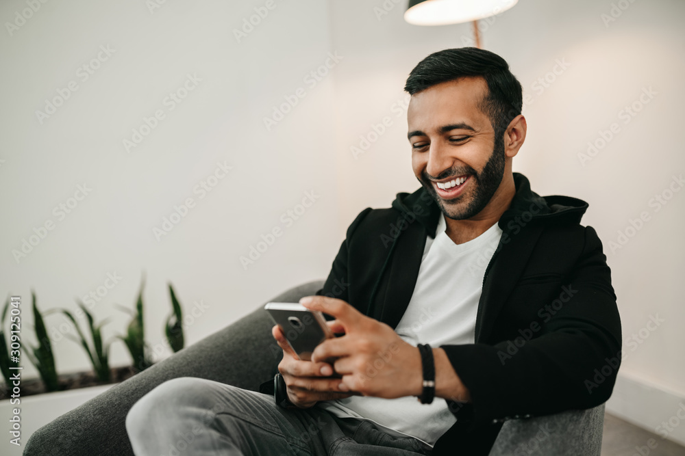 Trendy handsome Gen z Indian arabic man typing on mobile phone, smiling and laughing - obrazy, fototapety, plakaty 
