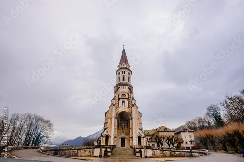 Medieval stone high catholic church on a round square Annecy France. Photography of a high stone tower with large arched windows on a background of snowy mountains, autumn landscape with cloudy sky.
