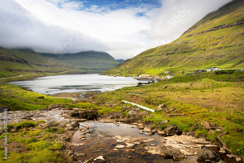 Lake mirror near small town and green mountains covered with fluffy clouds, Faroe Islands. © juhrozian