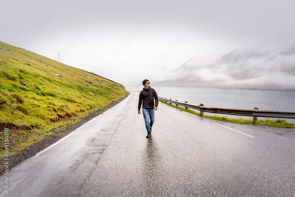 Young traveller walking y empty road after rain, green mountains, fjord and overcast sky on horizon. Faroe Islands, Denmark.