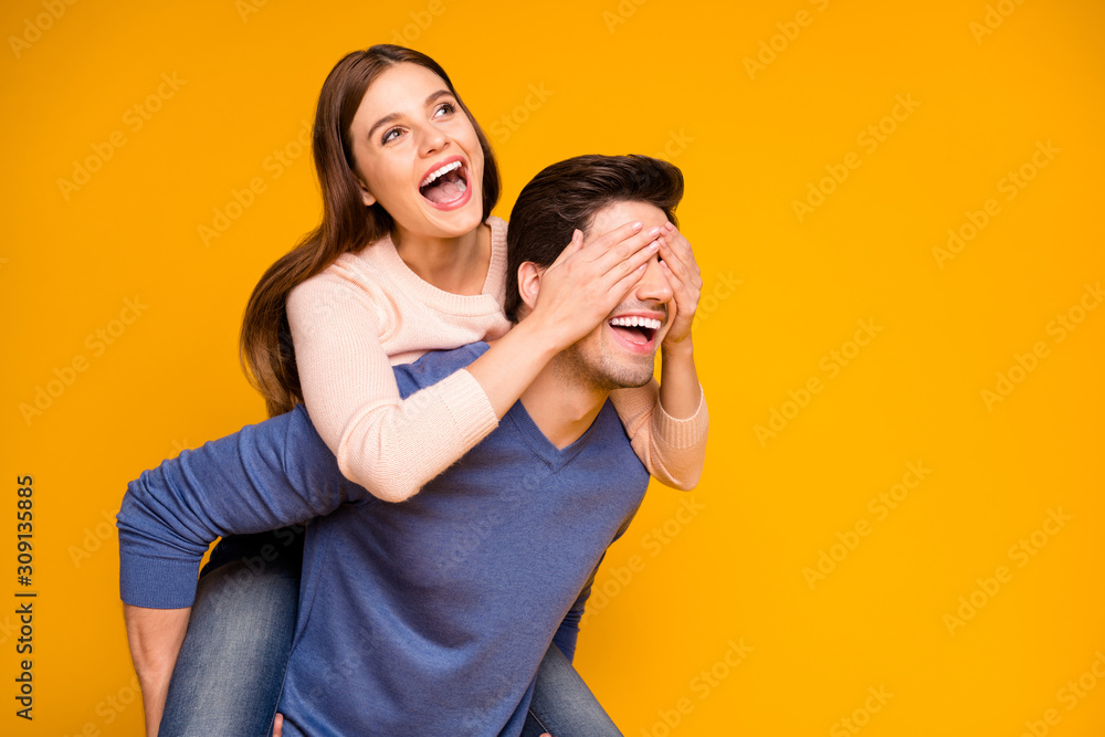 Positive cheerful woman close cover her spouse man eyes hug embrace piggy-back enjoy valentine day celebration wear blue pink sweater isolated over yellow color background Positive cheerful woman close cover her spouse man eyes hug embrace piggy-back enjoy valentine day celebration wear blue pink sweater isolated over yellow color background