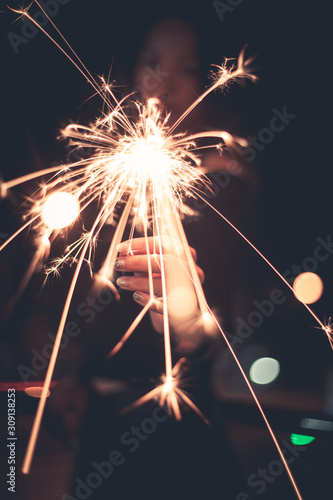 Lady holding sparklers stick on fire show forward, flame light spread out to camera at outdoor with light bokeh of light bulb at night time
