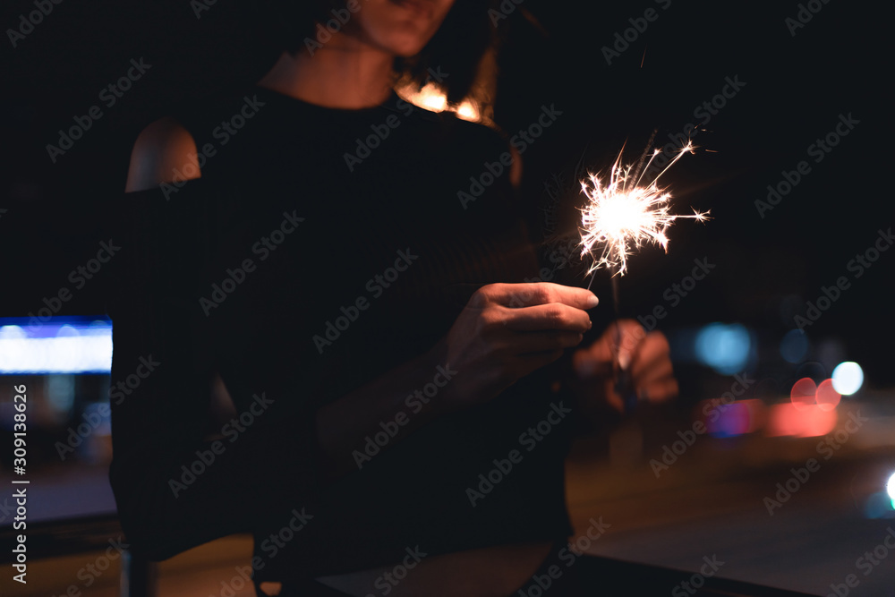 Lady holding both of sparklers sticks on fire to light up another one at outdoor with light bokeh of light bulb and car moving at night time Lady holding both of sparklers sticks on fire to light up another one at outdoor with light bokeh of light bulb and car moving at night time