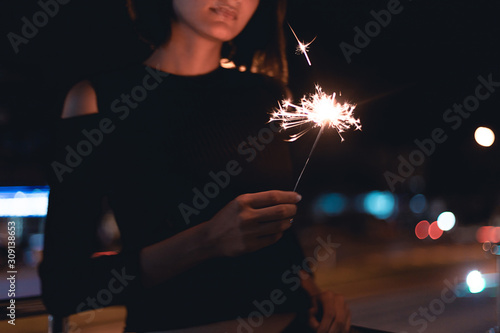 Lady holding sparklers stick on fire at outdoor with light bokeh of light bulb and car moving at night time