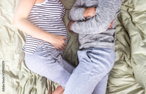 Young pregnant couple sleeping in bed on sunny morning. Sun rays shine through the window. Comfortable posture for sleeping. Top view. Minimalism lifestyle. Plain gren linens and gray clothes.