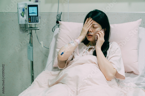 Sad disabled woman patient wearing nasal cannula talking on cellphone sitting in bed in hospital room. upset young girl in pajamas chatting with friend on smart phone using one hand cover face indoor photo