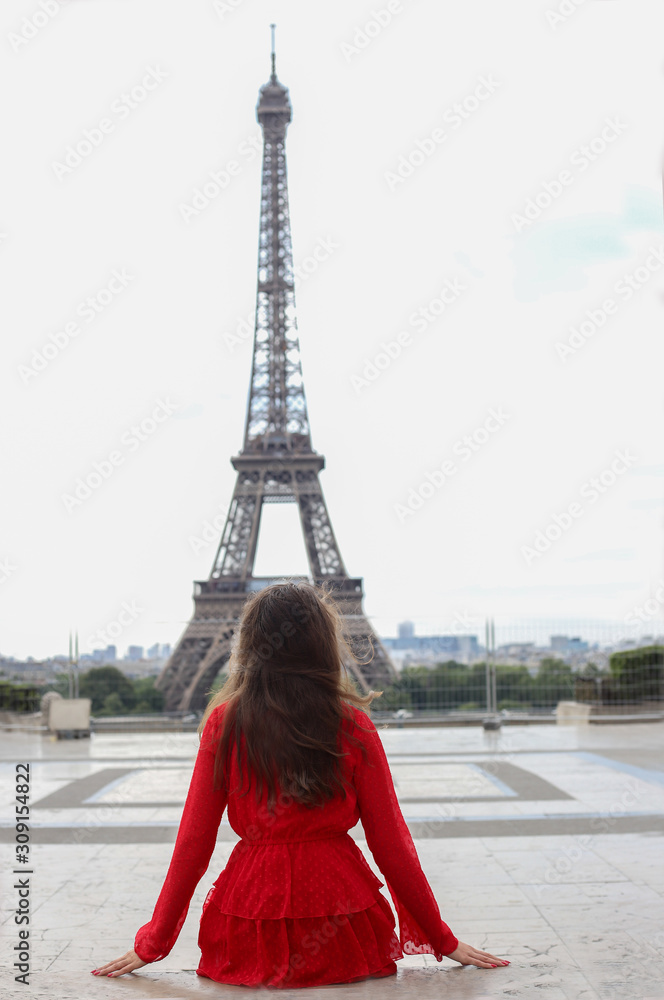 Young woman with long brown hair in red dress sitting back and look at Eiffel tower