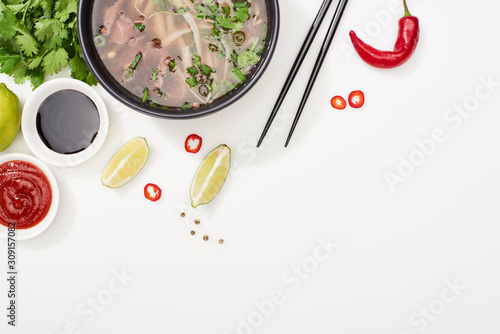 top view of pho in bowl near chopsticks, lime, chili and soy sauces and coriander on white background