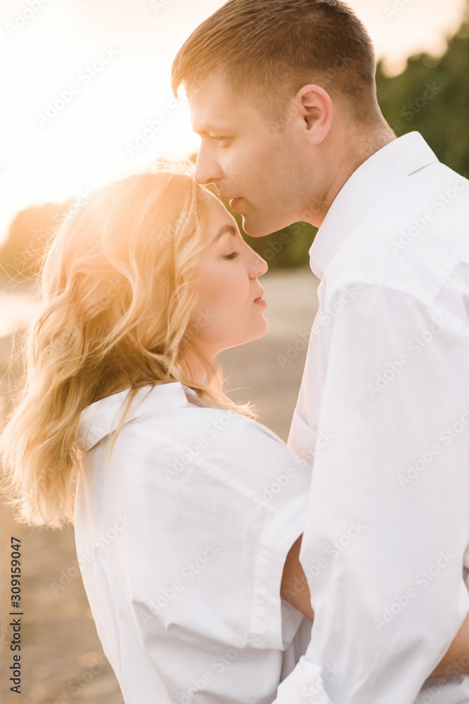 A young couple walks along the beach at sunset. The husband hugs his wife and kisses her forehead. The wife closed her eyes and smiles. Close-up. A young couple walks along the beach at sunset. The husband hugs his wife and kisses her forehead. The wife closed her eyes and smiles. Close-up.