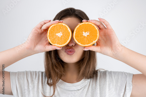 young brunette womanholding two halves of oranges near her eyes sending kiss standing on isolated white background dietology and nutrition