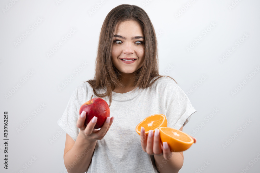 young attractive woman holding two halves of oranges in one hand and red apple in another looking surprised on isolated white background dietology and nutrition young attractive woman holding two halves of oranges in one hand and red apple in another looking surprised on isolated white background dietology and nutrition