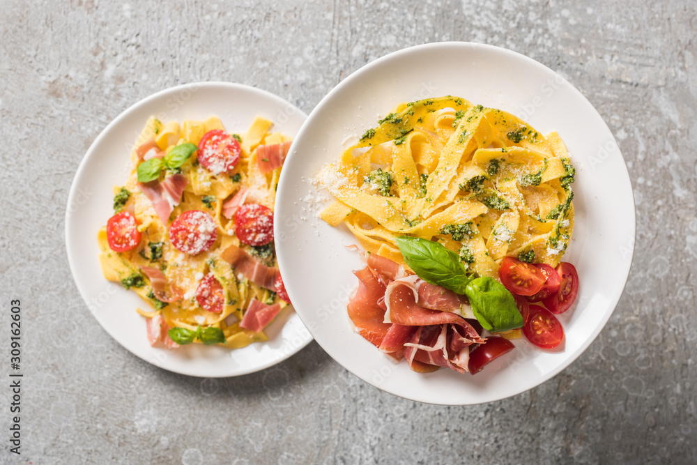selective focus of cooked Pappardelle with tomatoes, basil and prosciutto on plates on grey surface selective focus of cooked Pappardelle with tomatoes, basil and prosciutto on plates on grey surface