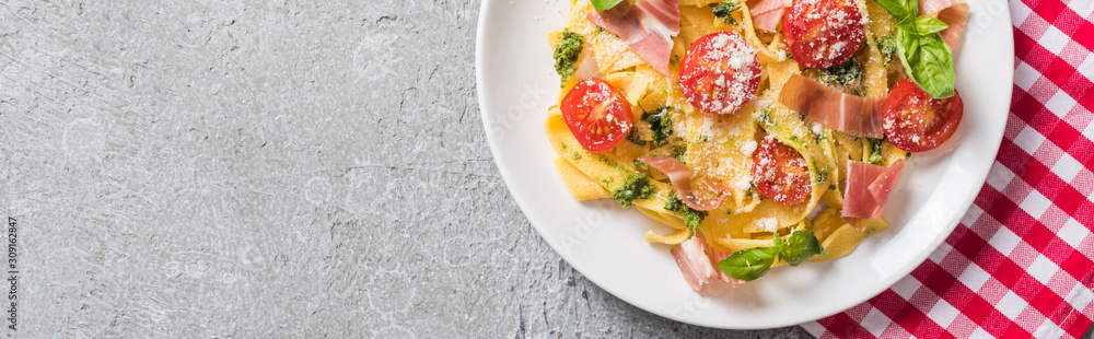 top view of cooked Pappardelle with tomatoes, basil and prosciutto on plaid napkin on grey surface, panoramic shot