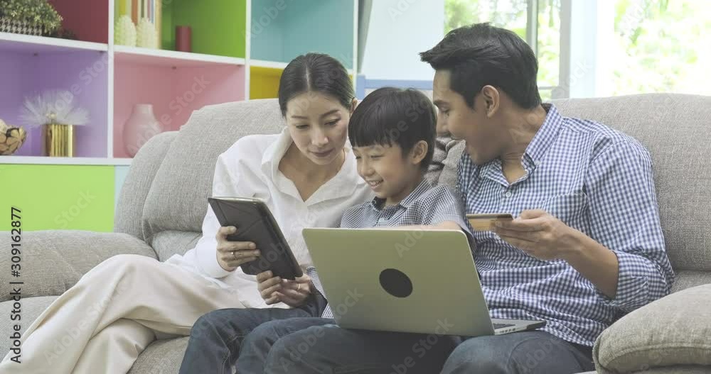 Family using laptop in living room. Asian man, woman and child sitting on couch. Man holding credit card, child key in the card. Family using laptop in living room. Asian man, woman and child sitting on couch. Man holding credit card, child key in the card.