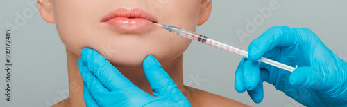 panoramic shot of beautician in latex gloves holding syringe near lips of woman isolated on grey