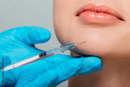 cropped view of beautician in latex gloves holding syringe near chin of girl isolated on grey