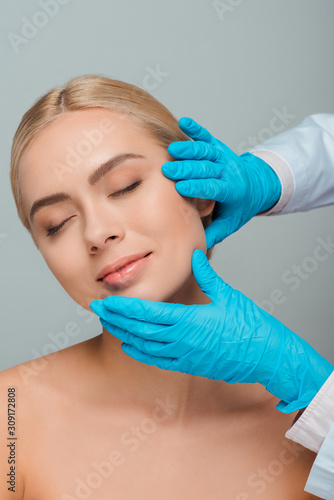 cropped view of beautician in latex gloves touching face of happy young woman isolated on grey