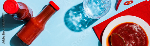 Top view of ketchup in plate and bottles beside chili pepper and glass of water on blue surface, panoramic shot
