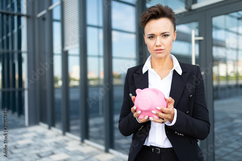 Caucasian woman in office clothes holds a pink pig moneybox and waits a coleague near the office building photo